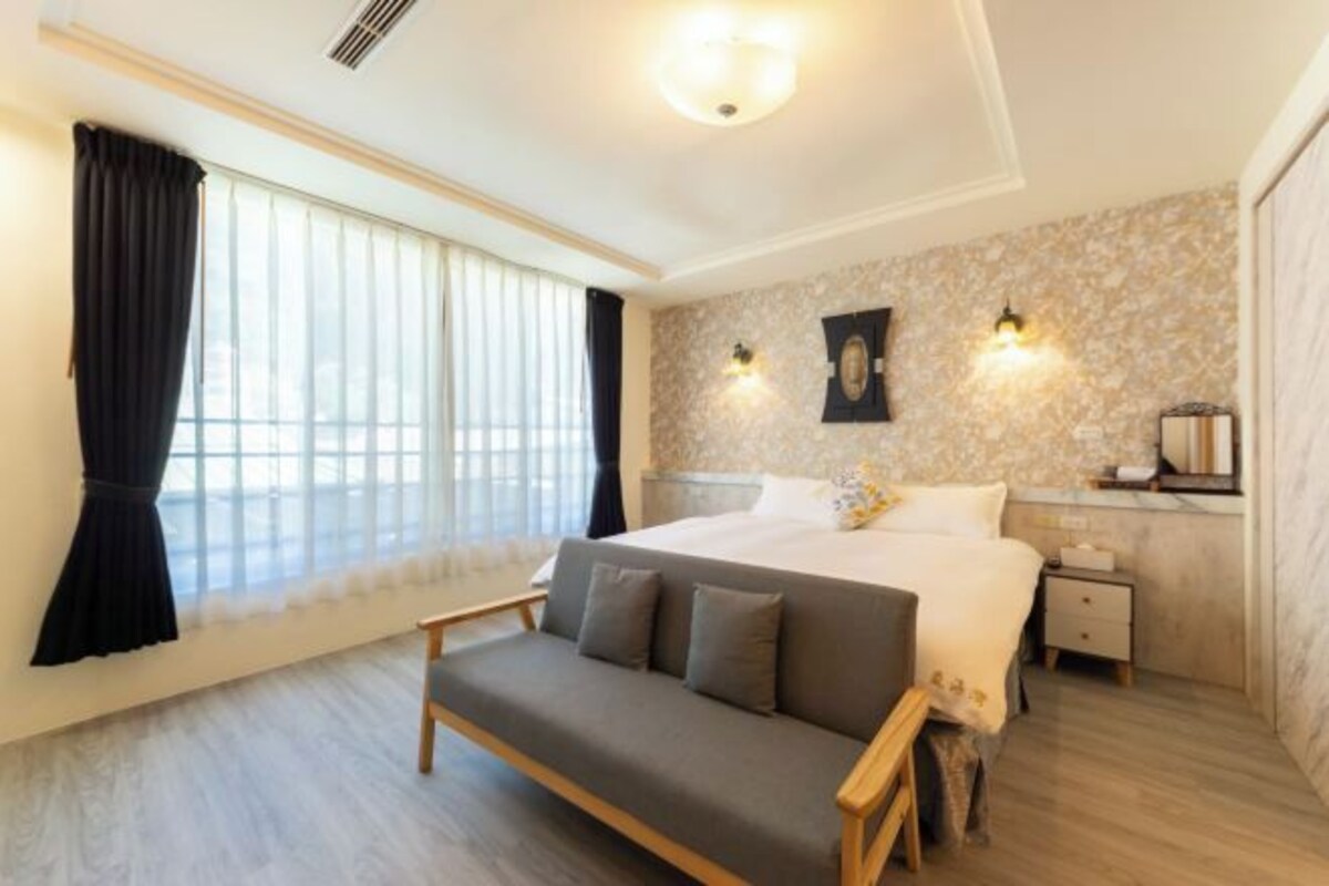 A bedroom with a large bed, a bedside table, a nightstand, a sofa, and a window with curtains.
