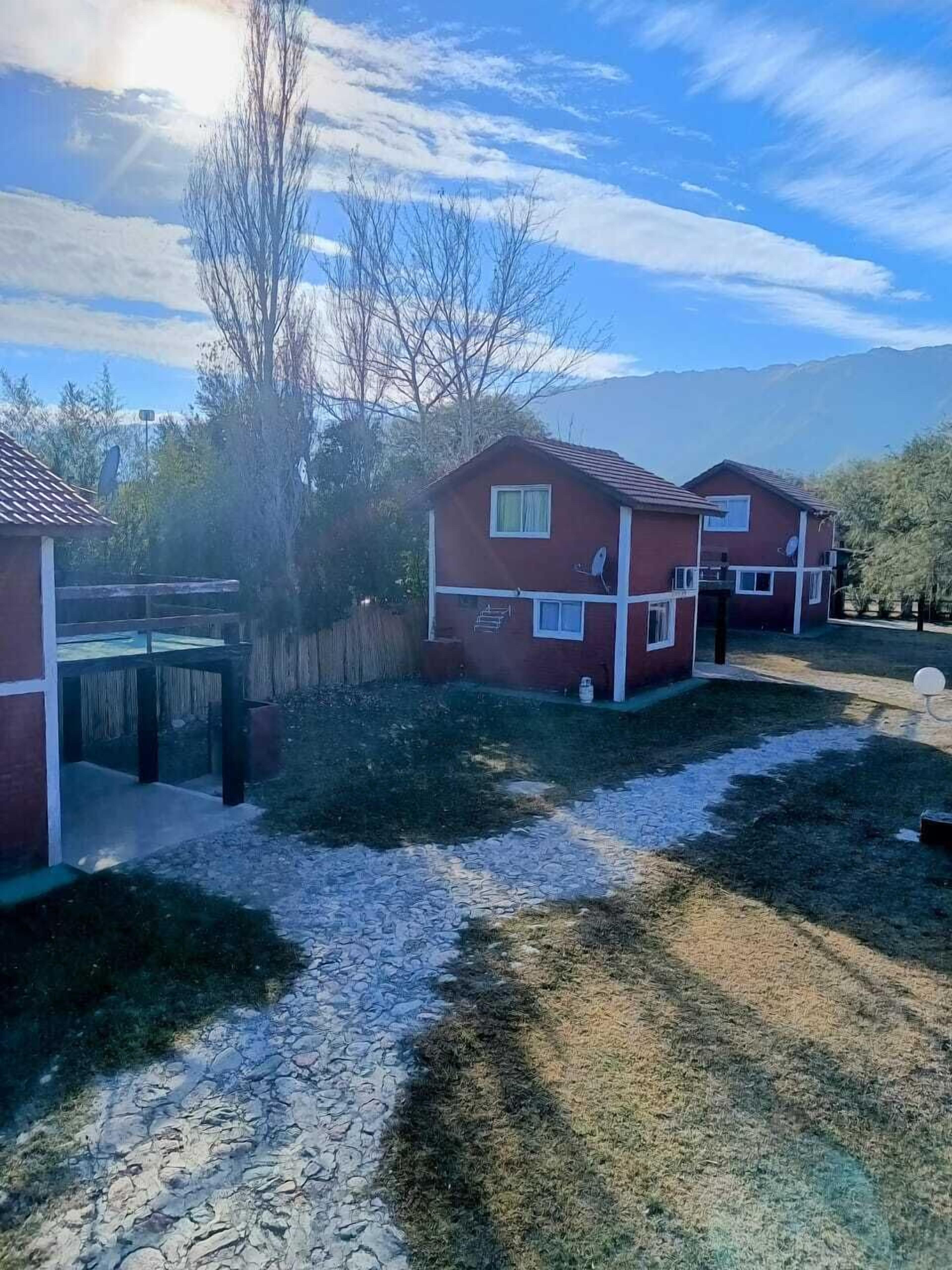 Um edifício de dois andares com fachada vermelha e detalhes brancos, um caminho de cascalho que leva à entrada, e um céu claro com montanhas ao fundo.