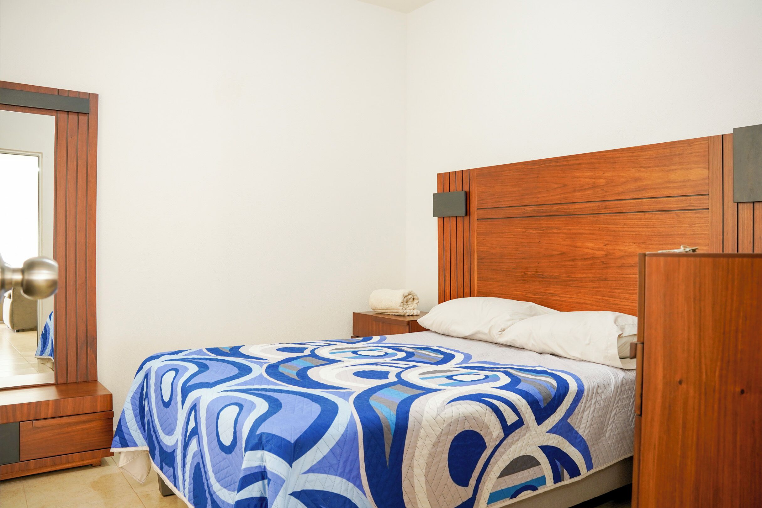 Un dormitorio con cabecera de madera, cama con colcha de diseño azul y blanco, mesita de noche con lámpara y espejo en la pared.