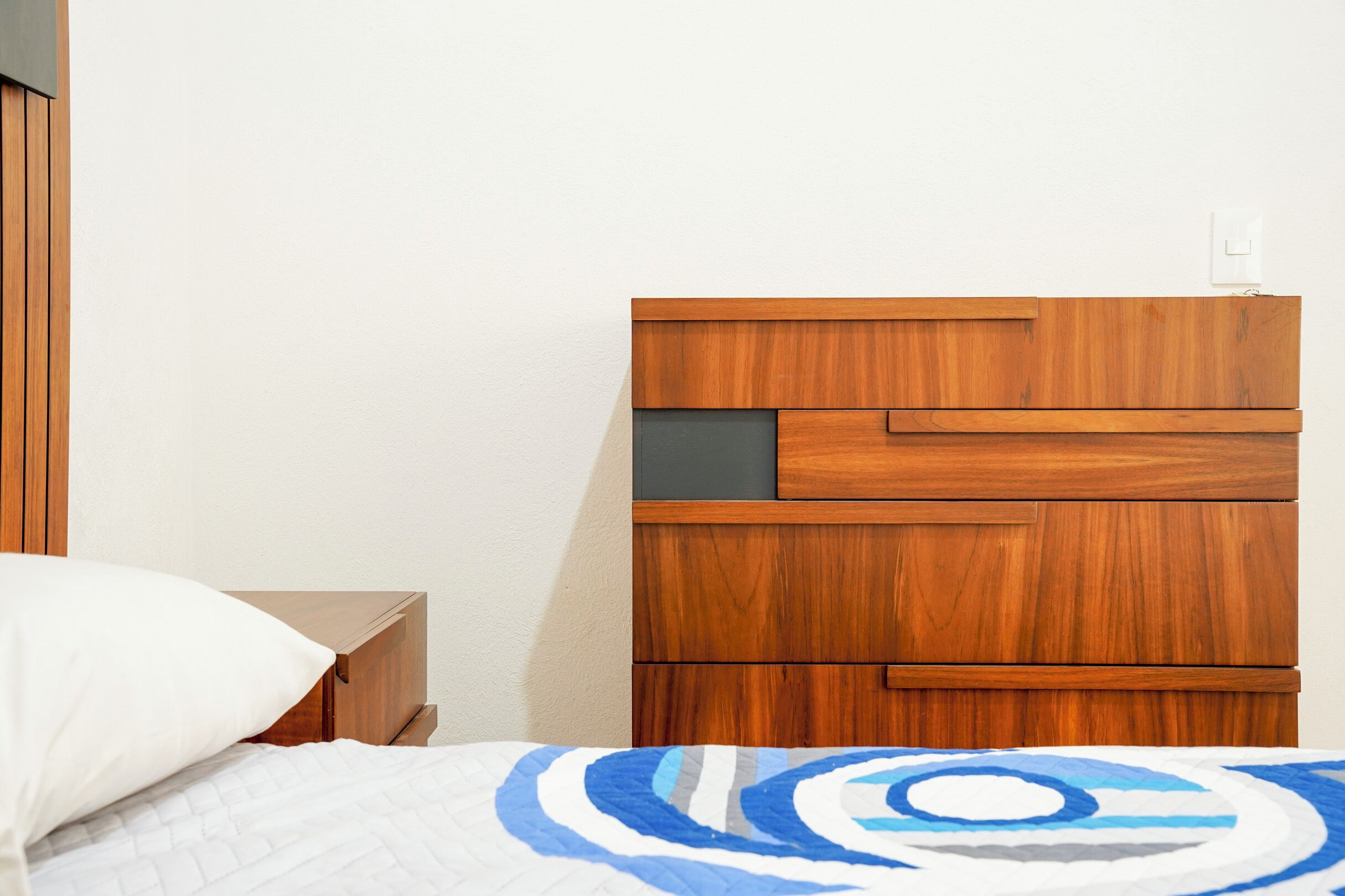 A bedroom with a wooden headboard, a bed with a white and blue patterned bedspread, and a nightstand with a white pillow.