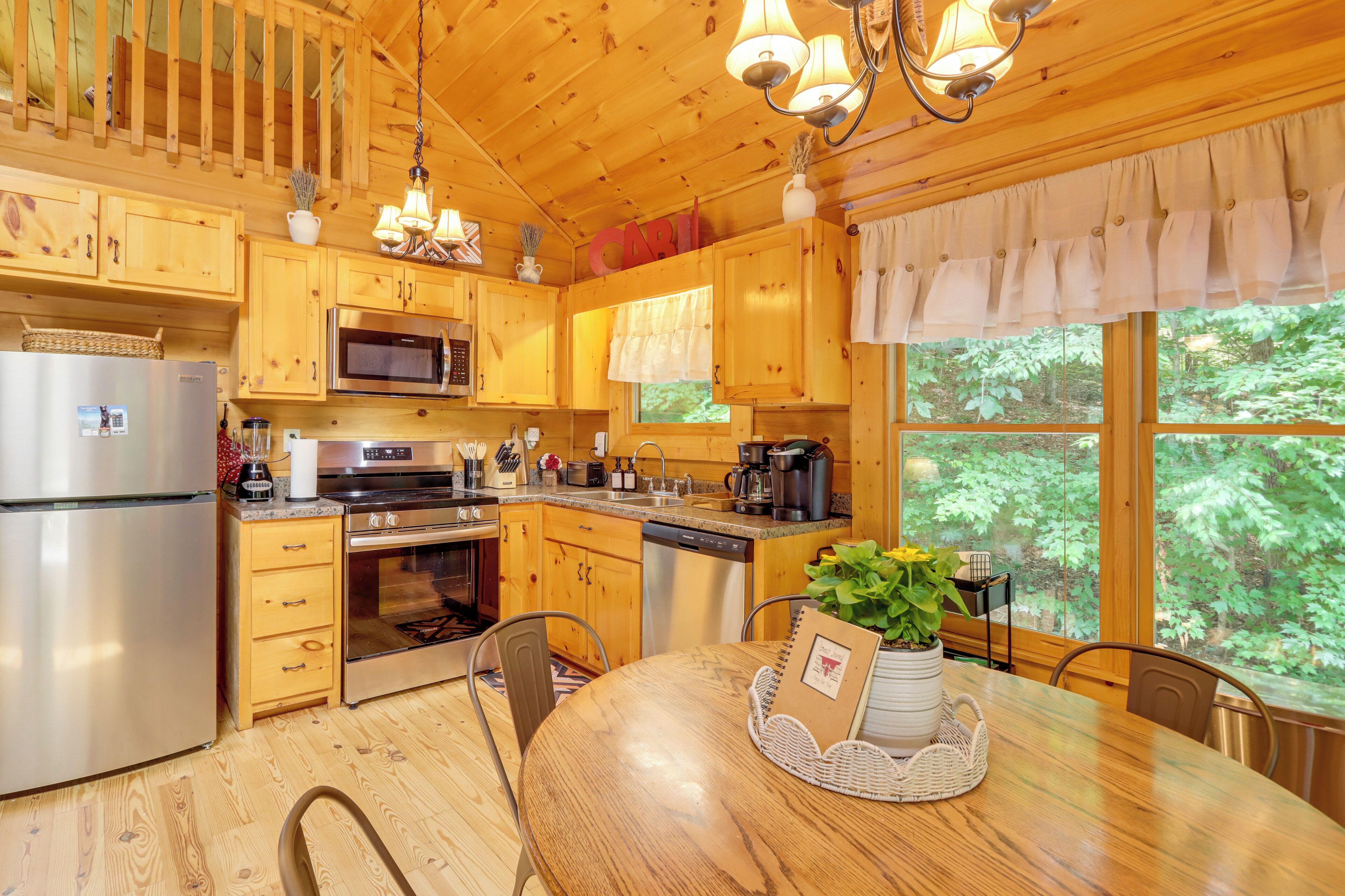 A wooden cabin kitchen with a dining table, wooden cabinets, stainless steel appliances, and a view of the forest.