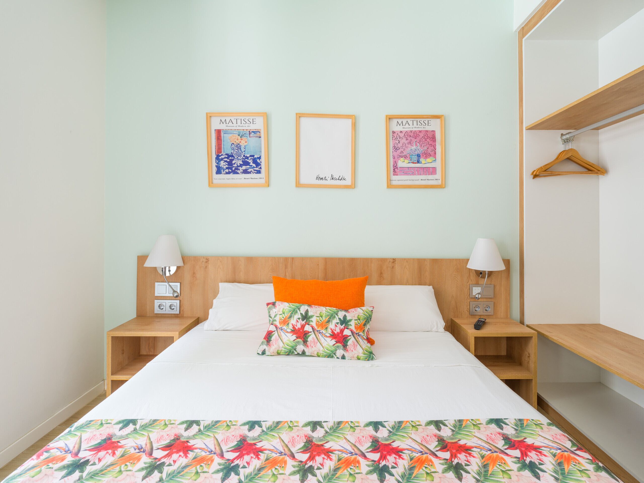 Una habitación de hotel con una cama, dos mesitas de noche, un armario y tres cuadros enmarcados en la pared.