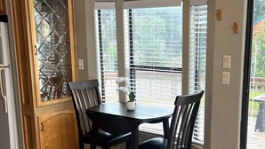 A dining area with a wooden table and chairs, a hanging light fixture, and a view through large windows. - The Rolling Rogue Cabin #49 (Gold Beach)