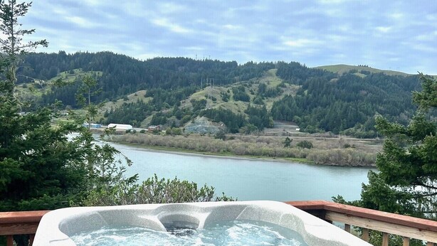 A hot tub on a deck with a view of a lake and hills. - The Rolling Rogue Cabin #49 (Gold Beach)
