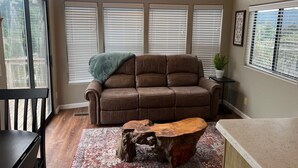 A cozy living room with a brown leather sofa, a wooden coffee table, and a large window with blinds. - The Rolling Rogue Cabin #49 (Gold Beach)