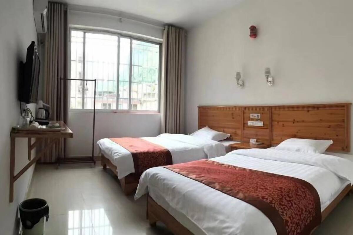 A hotel room with two beds, a desk, a chair, a trash bin, and a window with curtains.