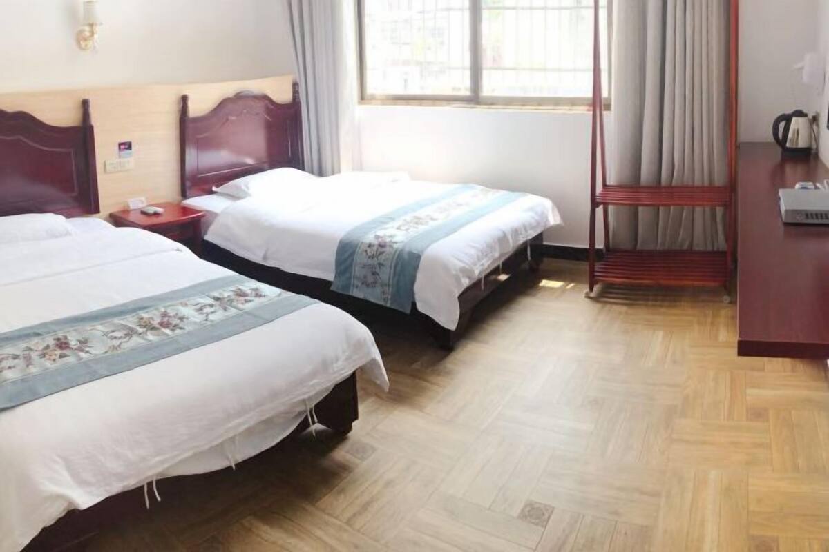 A hotel room with two beds, wooden flooring, a window with curtains, a nightstand, and a desk with a kettle.