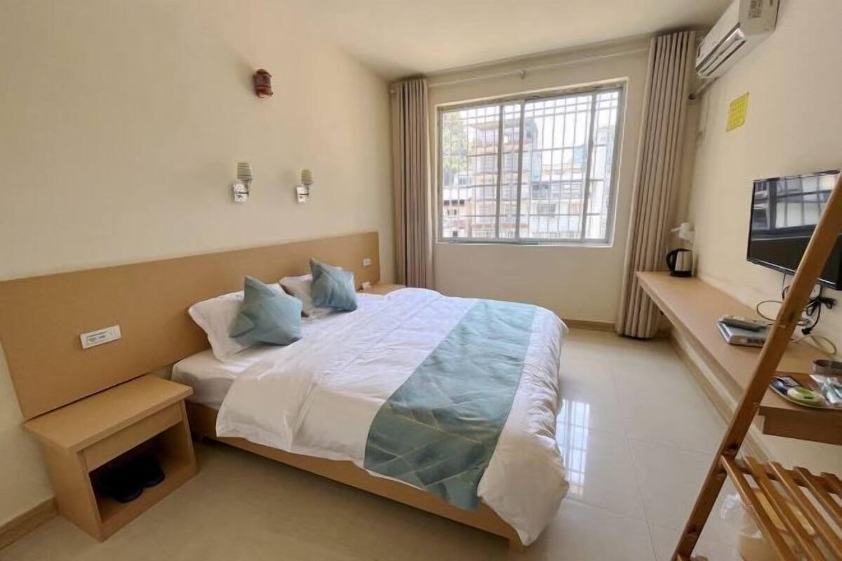 A hotel room with a bed, a nightstand, a desk with a chair, a TV, and a window with curtains.
