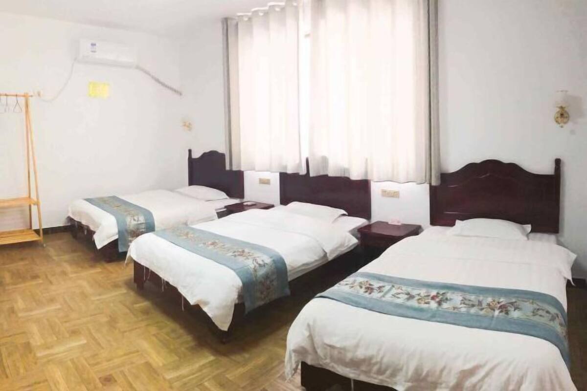 A hotel room with three single beds, a wooden floor, a window with curtains, and a wall-mounted air conditioner.