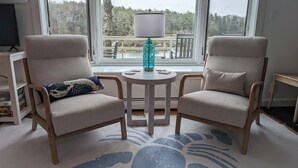 A room with two armchairs, a small table, and a lamp. There is a window with a view of trees and a deck outside.