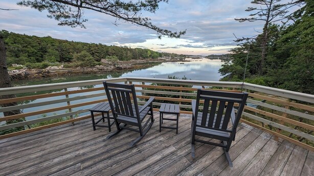 A wooden deck with two rocking chairs and a small table, overlooking a calm lake surrounded by trees.