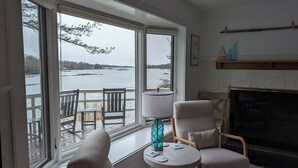 A cozy room with a large window overlooking a body of water, a fireplace, and a small round table with a lamp.