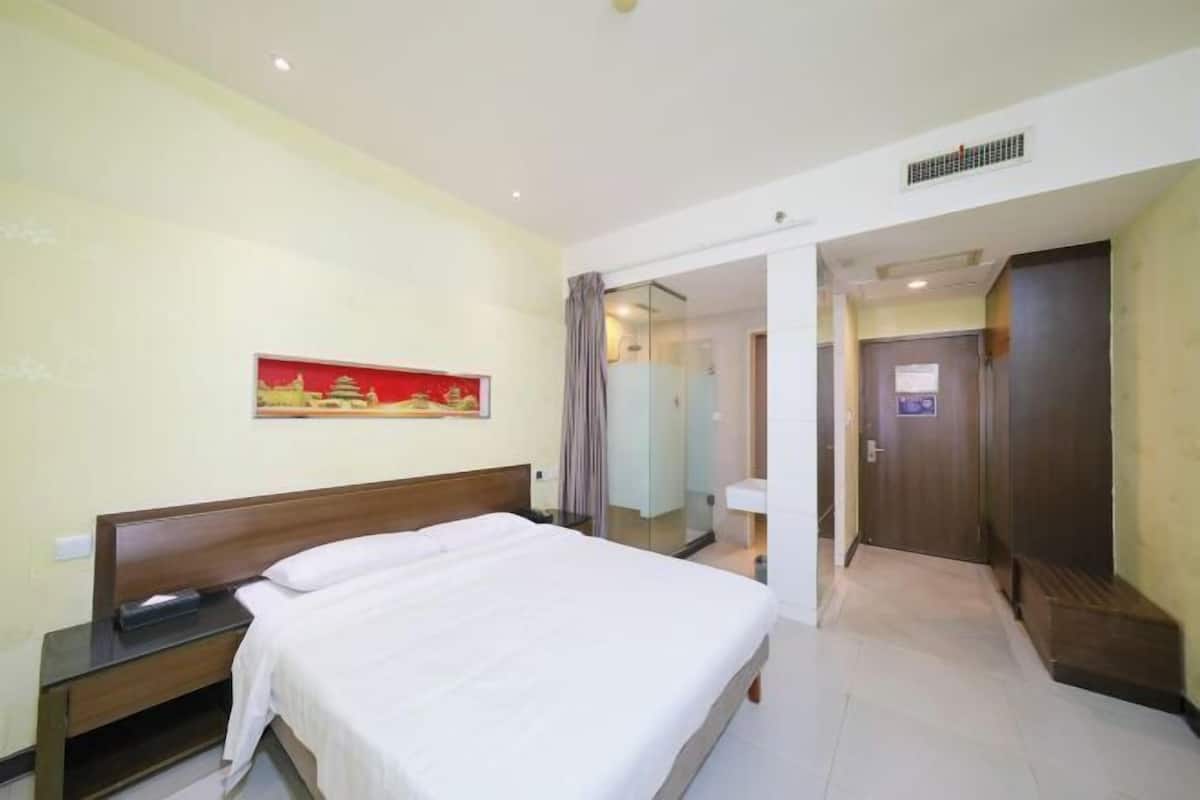 A hotel room with a bed, a nightstand, a wall-mounted painting, a bathroom with a mirror, and a door leading to another room.