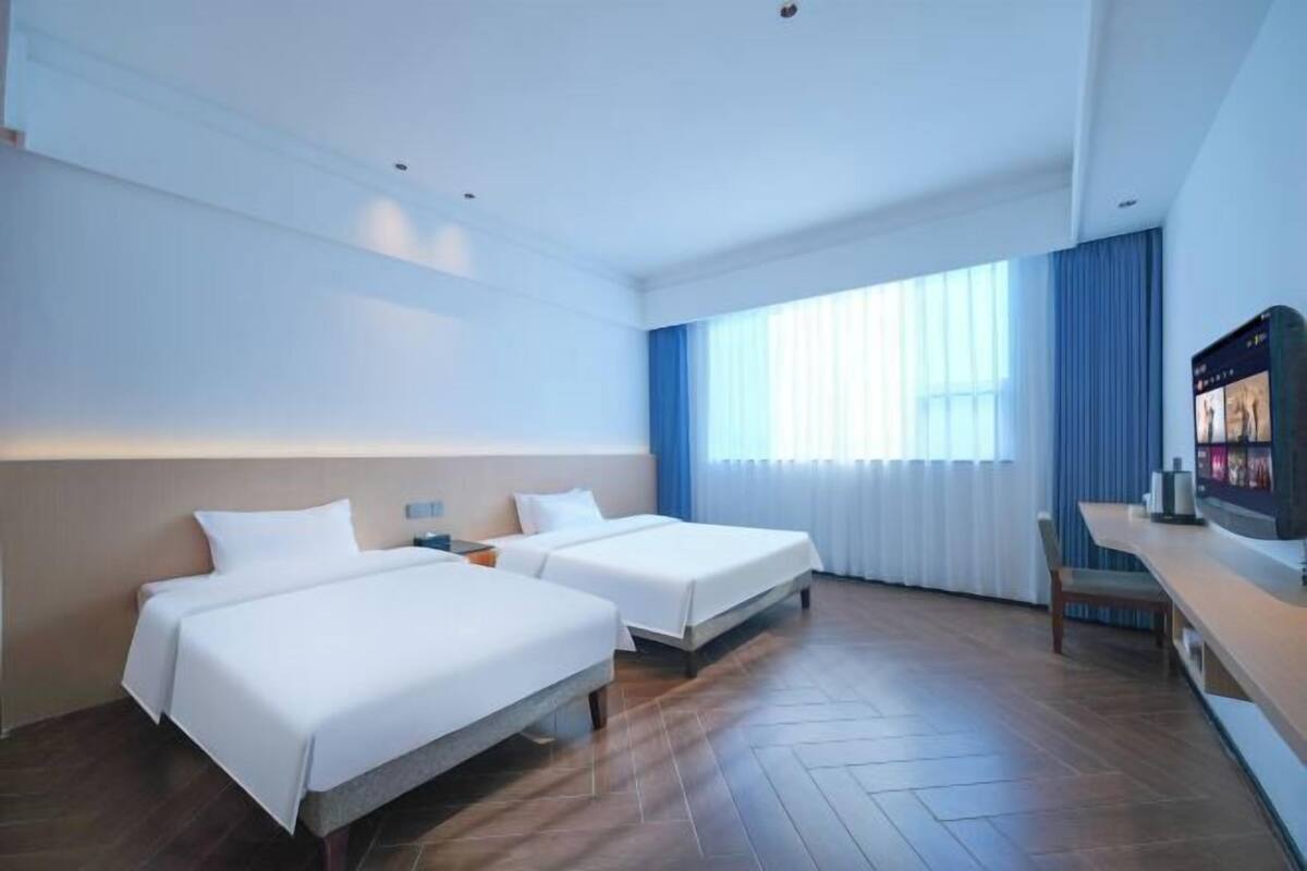 A hotel room with two beds, a desk, a TV, and a large window with curtains.