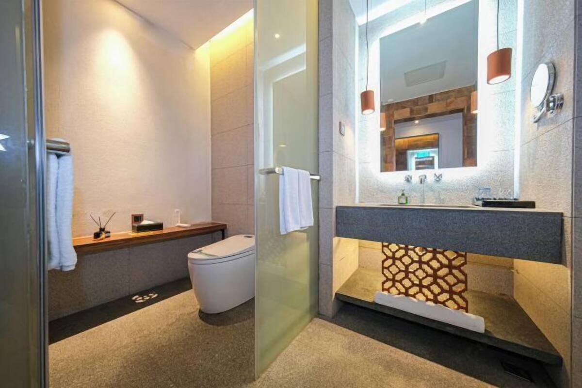 A modern bathroom with a glass shower enclosure, a toilet, a sink with a stone backsplash, and a mirror with pendant lights.