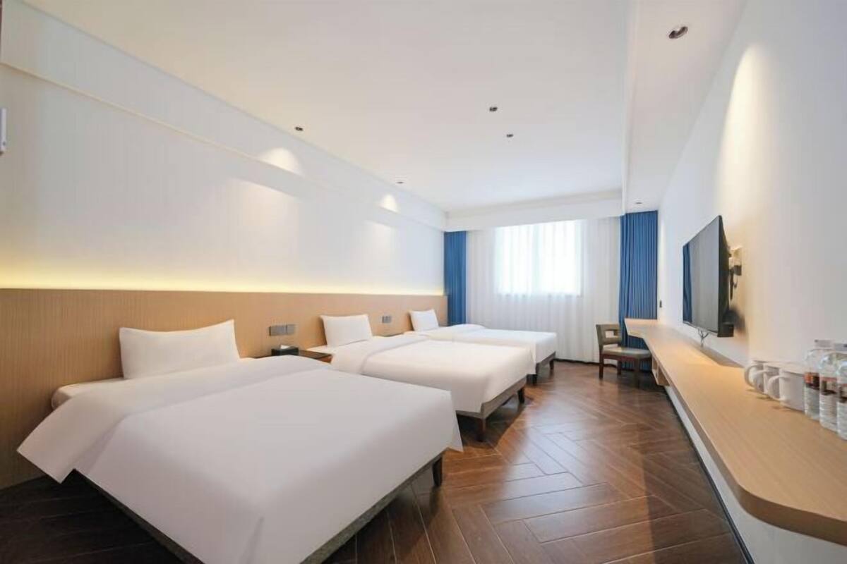 A hotel room with two beds, a wooden floor, a flat-screen TV, a minibar, and a window with curtains.