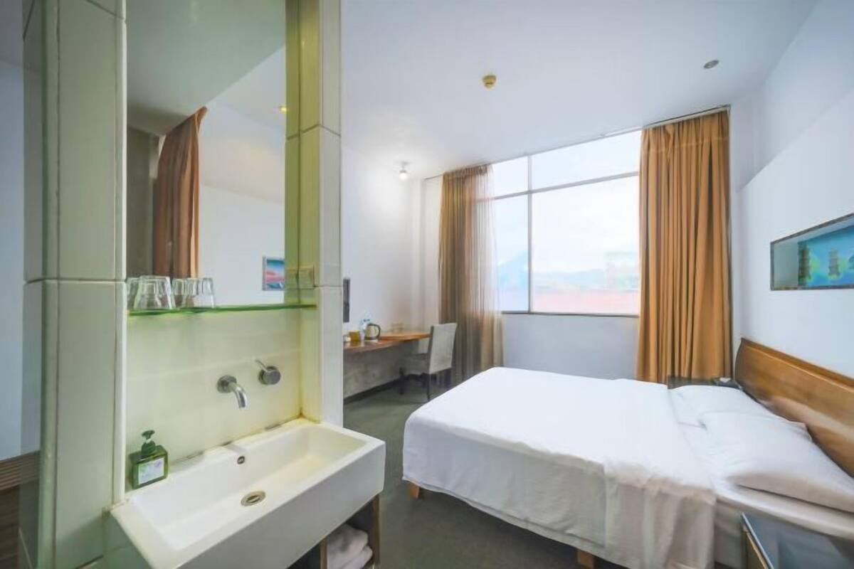 A hotel room with a bed, a sink, a mirror, a desk, and a window with curtains.