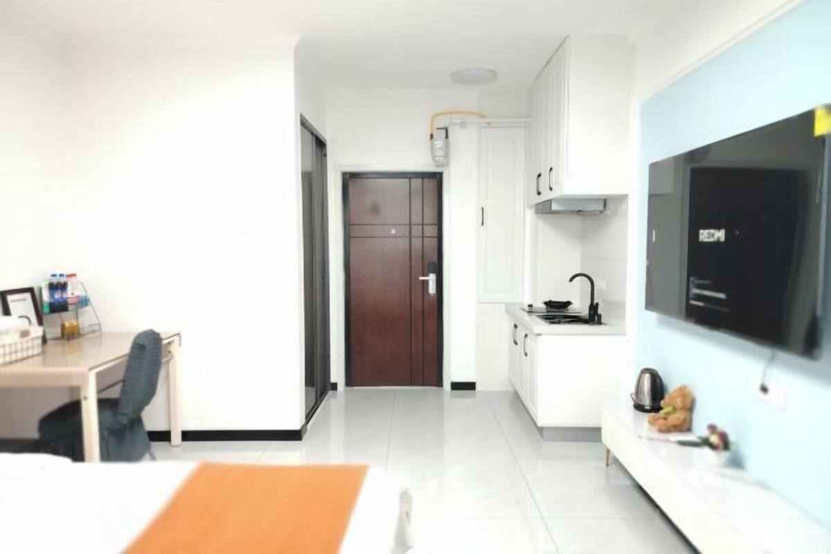 A modern hotel room with a bed, a desk, a kitchenette, and a TV.