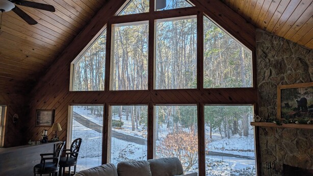 L’intérieur chaleureux d’un chalet, avec une grande fenêtre donnant sur un paysage enneigé, une cheminée et un ventilateur de plafond.