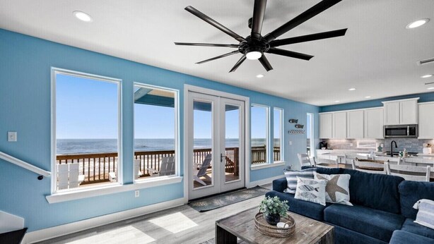A spacious living room with a large window overlooking the beach, a ceiling fan, and a kitchen area. - Fishy Business| Beach Front Oasis!  Sleeps 20! (Bolivar Peninsula)