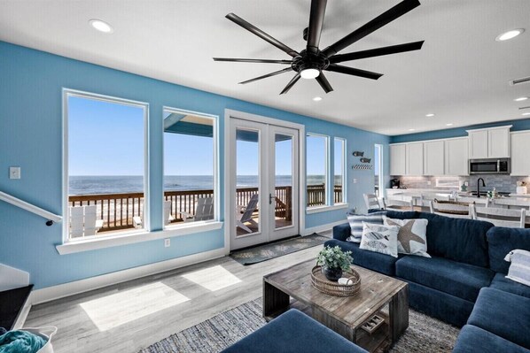 A spacious living room with a large window overlooking the beach, a ceiling fan, and a kitchen area.