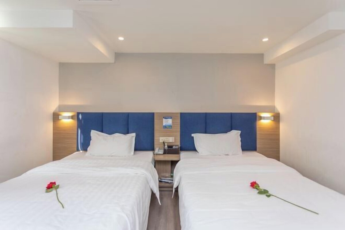 A hotel room with two beds, each with a wooden headboard and white bedding. There are bedside tables with lamps and a small clock.