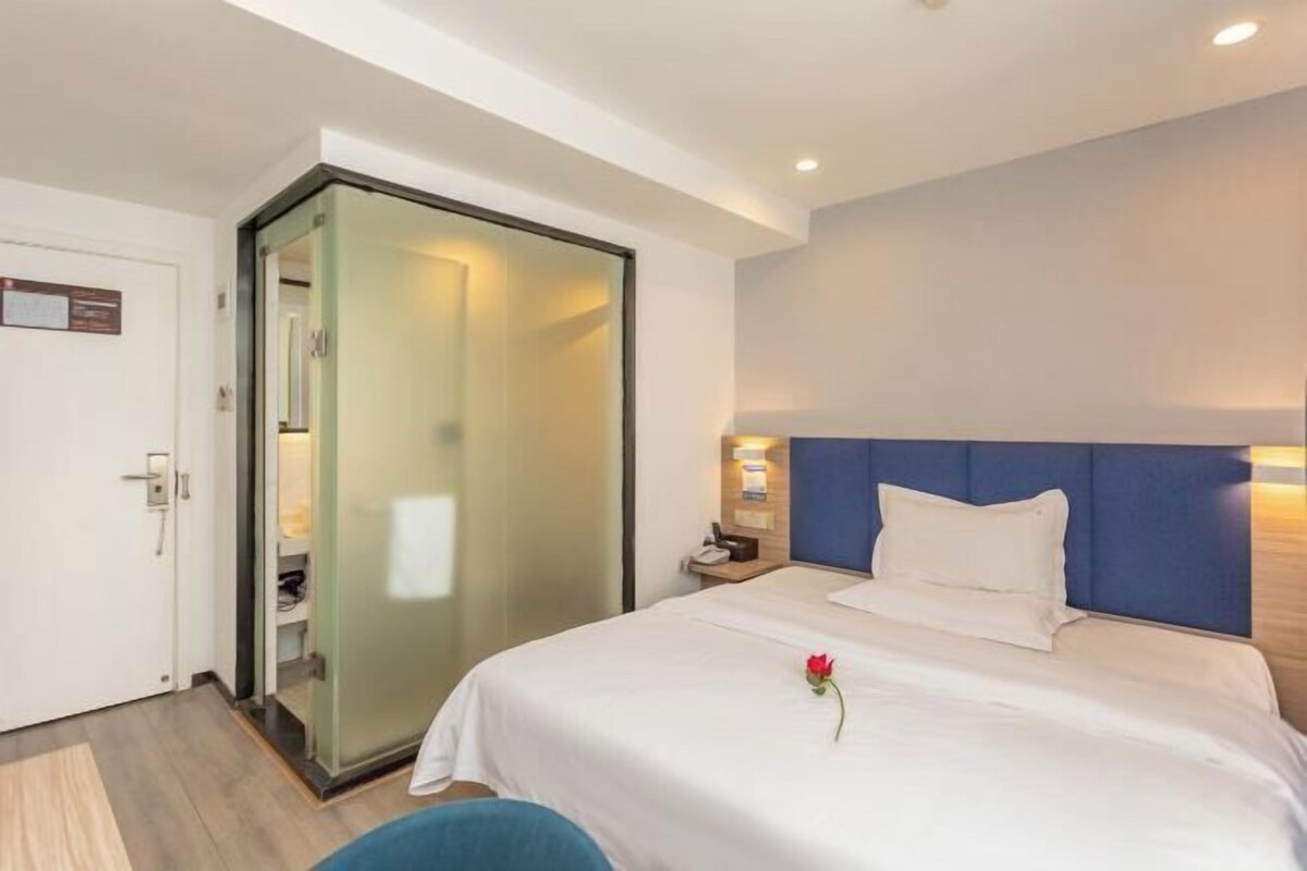 A hotel room with a bed, a nightstand, a blue headboard, and a glass door leading to a bathroom.