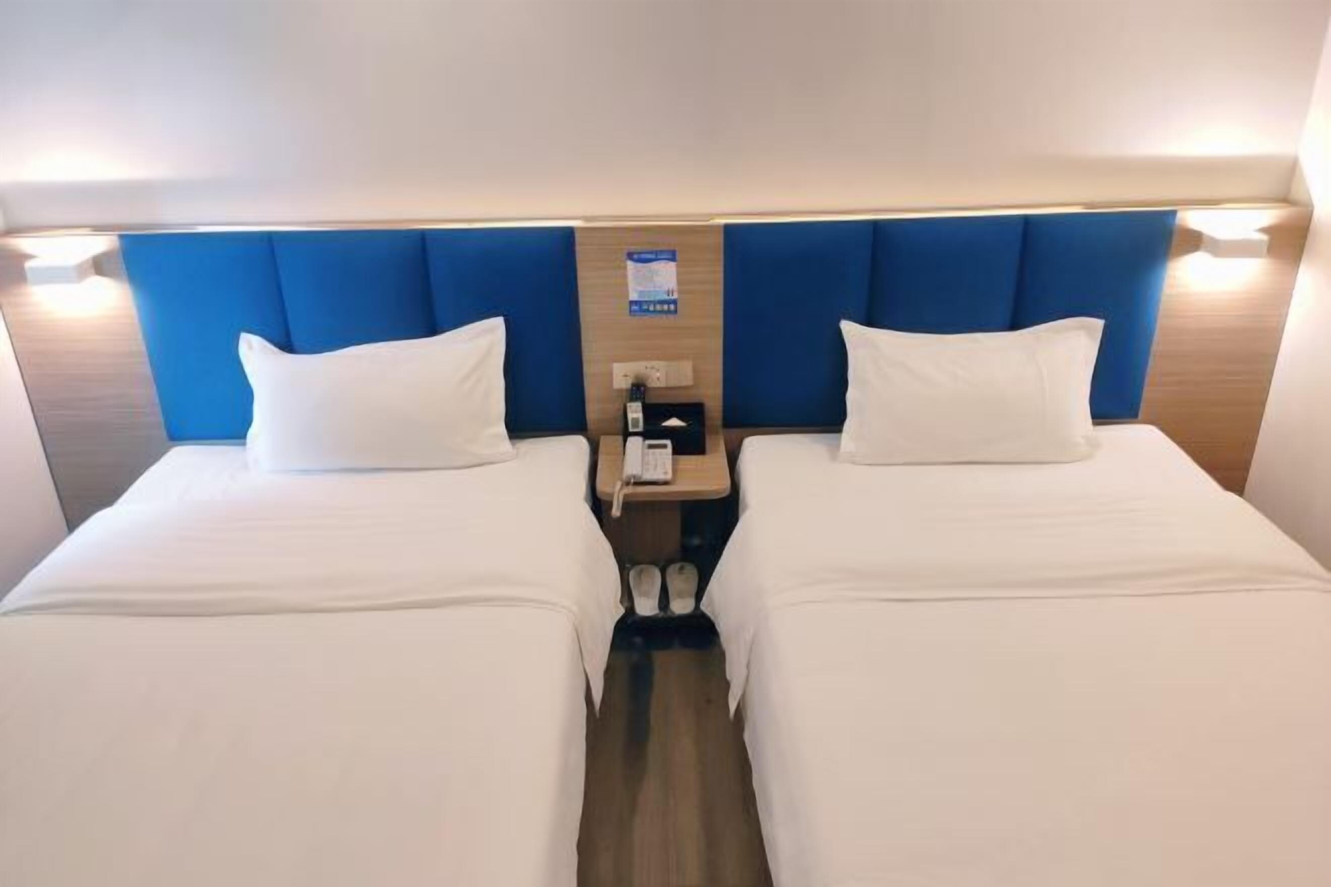 A hotel room with two beds, a wooden headboard, a nightstand with a phone, and slippers.