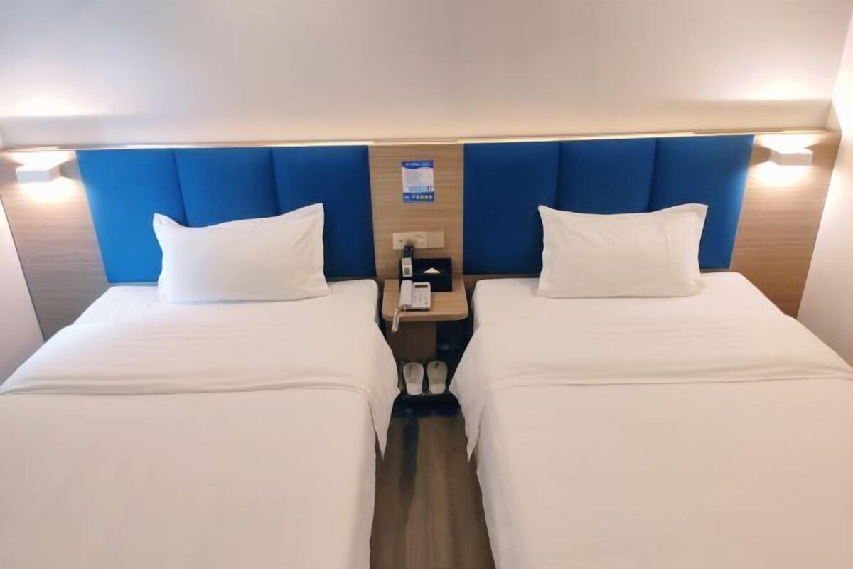 A hotel room with two beds, a wooden headboard, a nightstand with a phone, and slippers.
