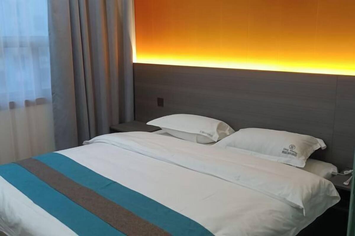 Una habitación de hotel con cama, ropa de cama blanca, cabecera con luz cálida ambiental y una ventana con cortinas translúcidas.