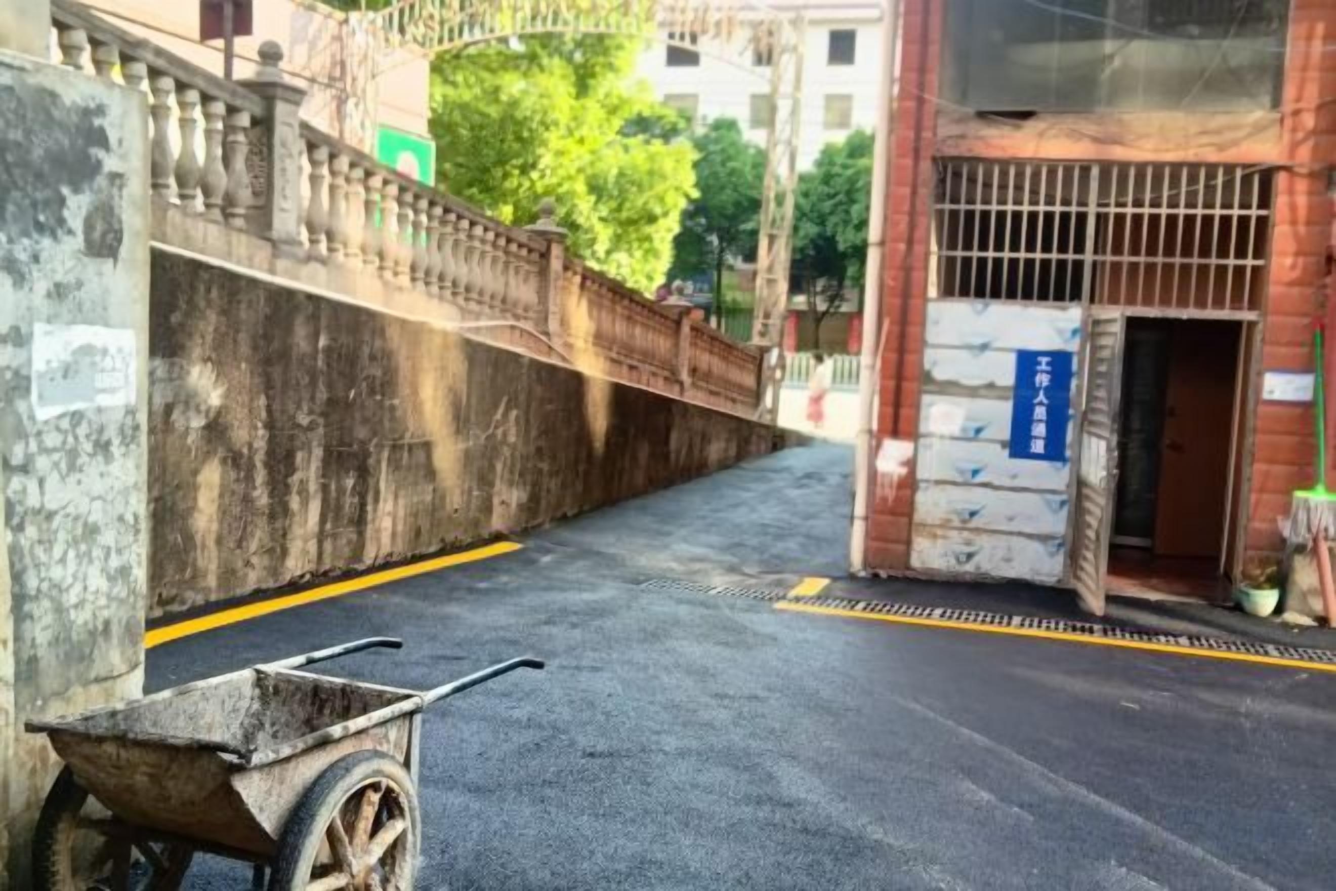 A street with a wheelbarrow, a concrete wall, and a building with a barred entrance.