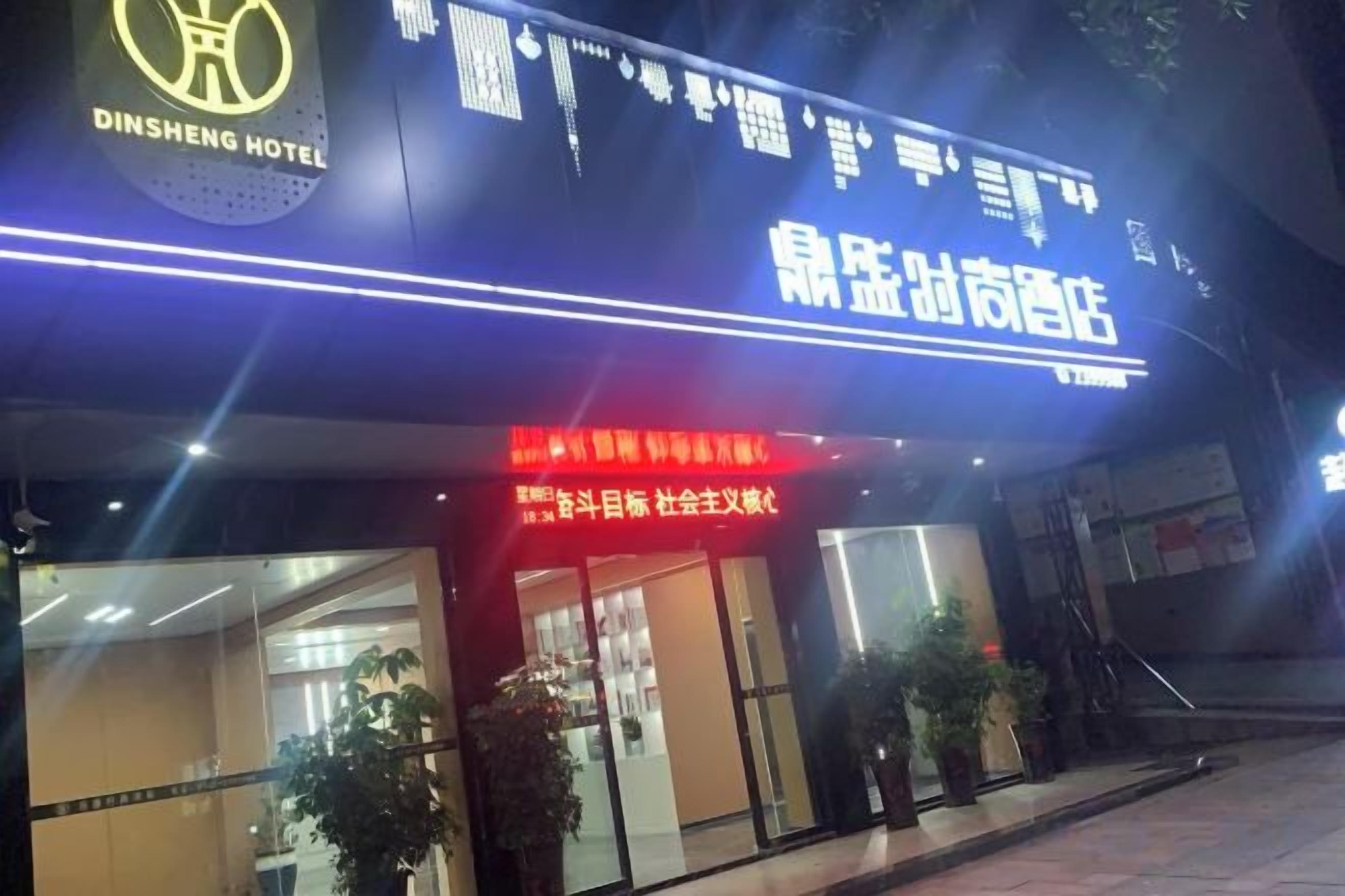 A hotel entrance with signage in a foreign language, a visible reception area, and potted plants outside.
