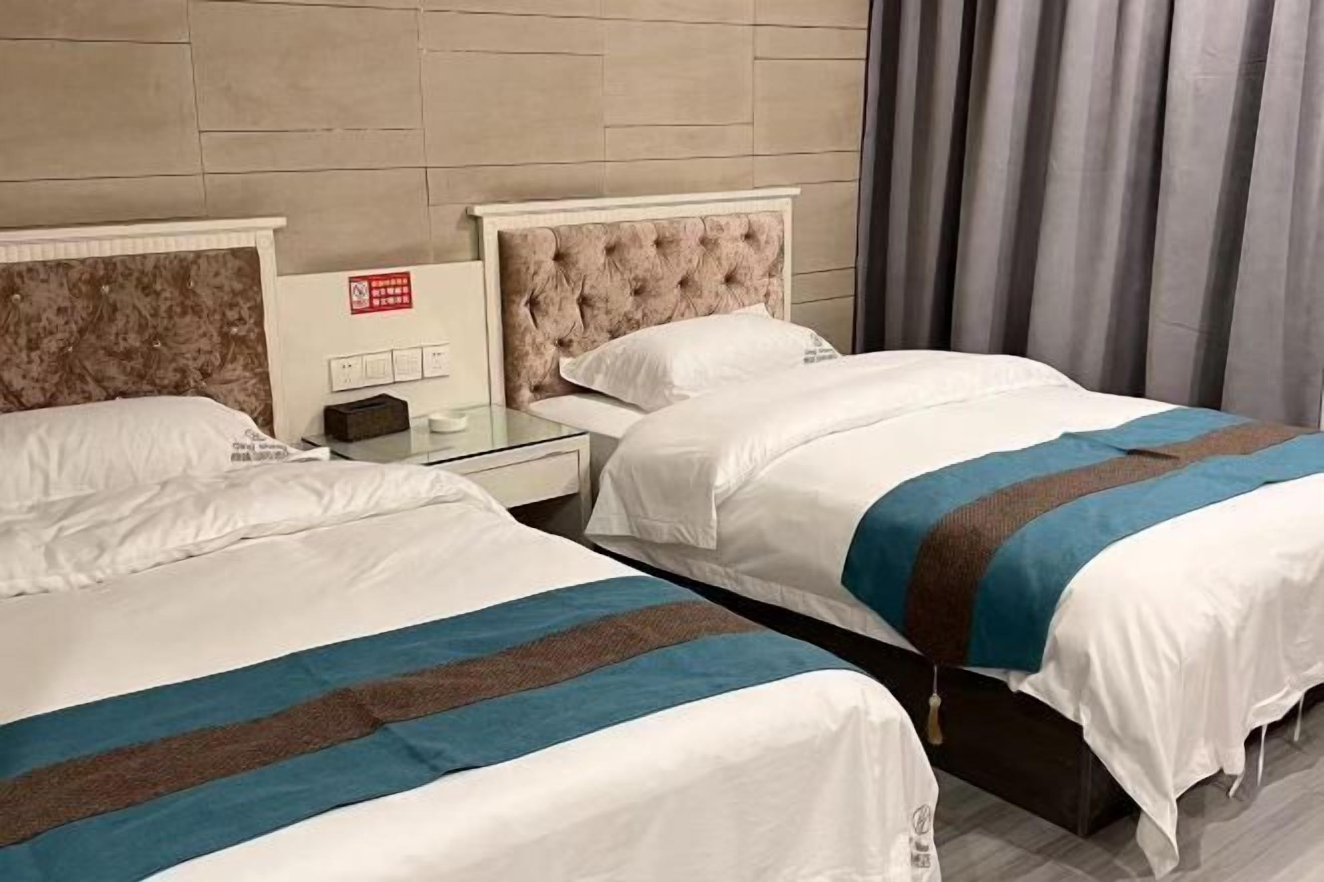 Twee bedden met witte beddengoed en blauwe strepen, een nachtkastje met een lamp, en een rookmelder aan de muur.
