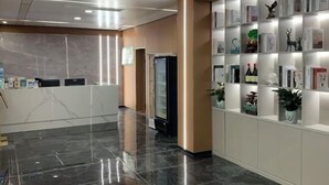 En hotellobby med en receptionsdisk, ett kylskåp och en vägg med hyllor där böcker och dekorativa föremål visas upp.