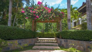 Un chemin de pierres mène à une pergola en bois, ornée de fleurs roses et entourée d’une végétation luxuriante et de palmiers.