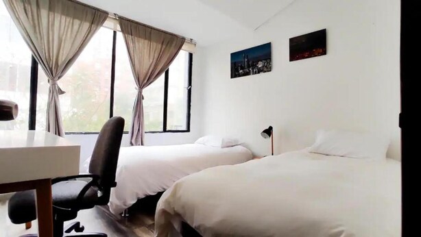 A hotel room with two single beds, a desk with a chair, and a window with curtains. - Apartamentos El Árbol (Bogotá)
