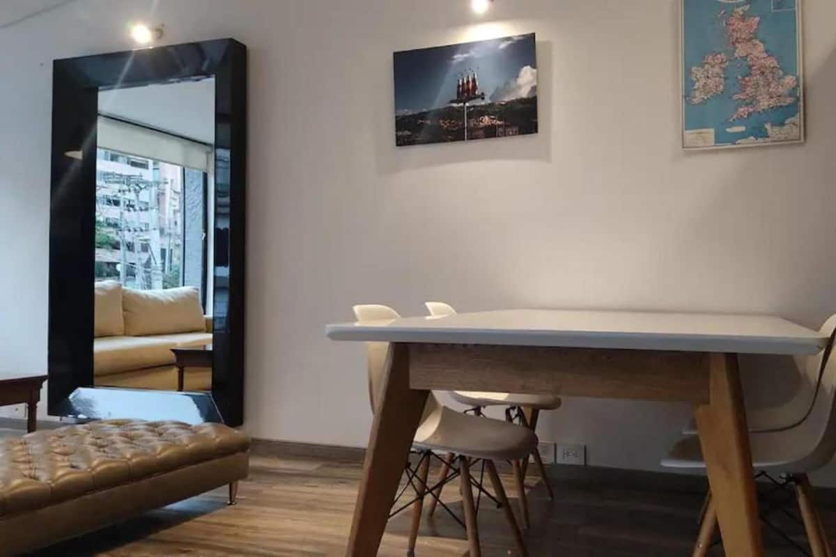 A modern room with a large mirror, a dining table with chairs, a sofa, and wall art including a map and a landscape painting.
