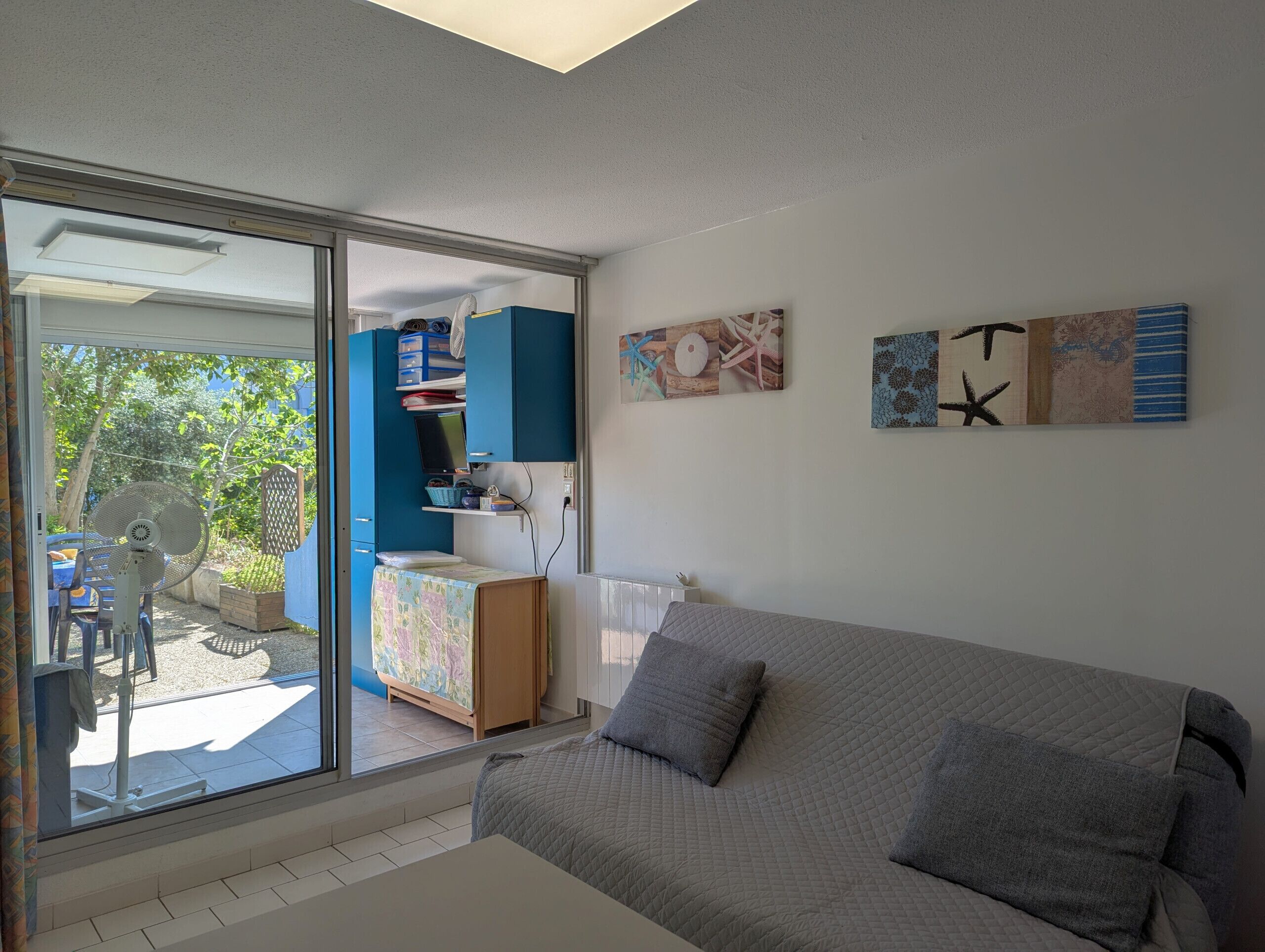 A modern living room with a grey sofa, a large sliding glass door leading to an outdoor area, and a blue cabinet with various items.