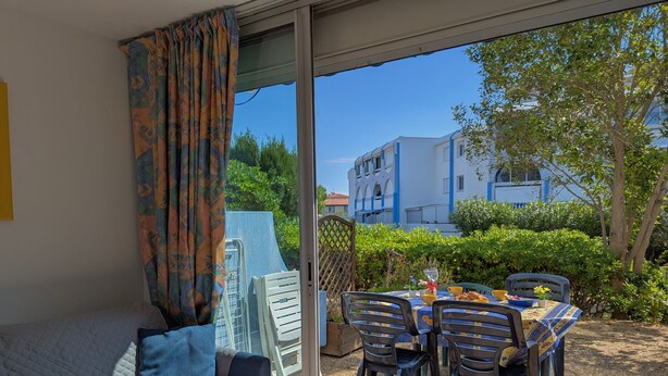 A room with a bed, a table with chairs, and a view of buildings and greenery outside. - Sol-y-Days Floralies 2, Studio with terrace and swimming pool (Le Grau-du-Roi)