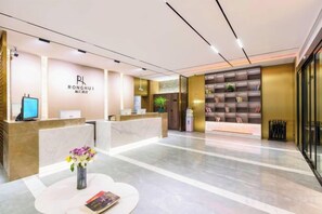 A hotel reception area with a reception desk, a seating area with a vase of flowers, and a wall with a plant and a shelf with books. - Korla Ronghui Yi Hotel (Xiangli Avenue Food Street Branch) (Korla)