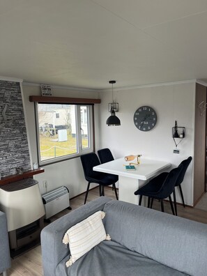 A modern living room with a white sofa, a dining table with two chairs, a black pendant light, a wall clock, and a stone wall.