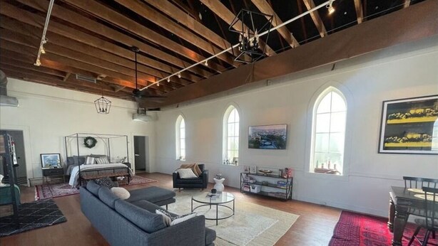 A spacious living room with a large gray sofa, a coffee table, and a dining area with a table and chairs. There are arched windows and a wooden ceiling. - St. Chris - Graham, TX (Graham)