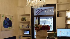 A modern hotel room with a chandelier, a sofa, a desk with a computer, and a view of a hotel building. - HOTEL ARISTON (Piombino)