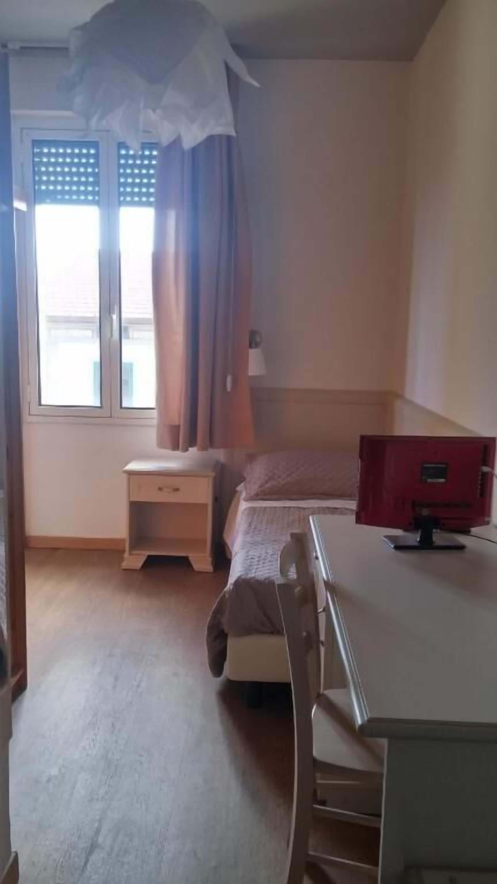Una habitación pequeña y bien iluminada con una cama, un escritorio y una silla. Cuenta con una ventana con cortinas, una mesita de noche y un armario rojo.