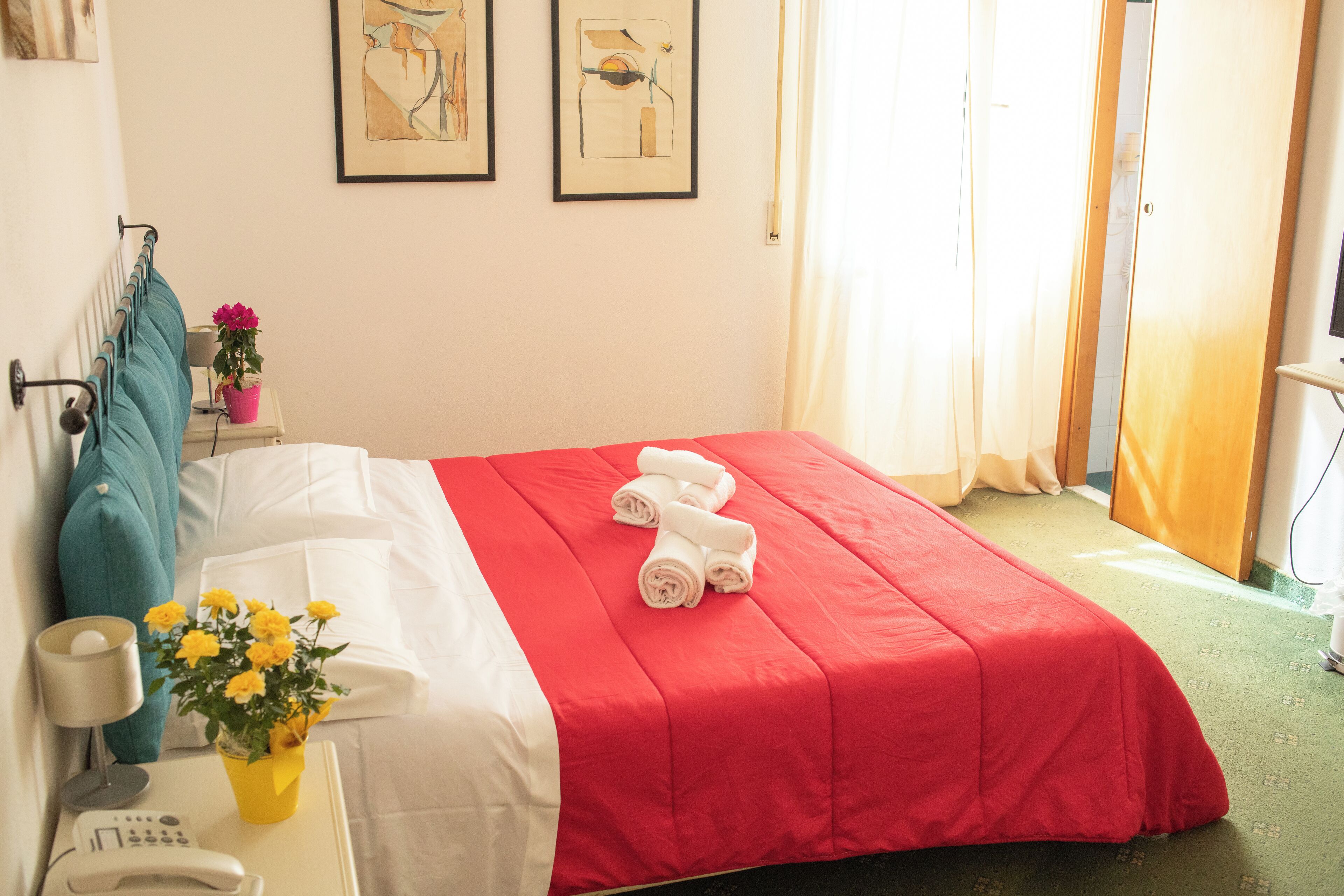 Une chambre d’hôtel avec un lit bien fait, une table de chevet avec une lampe et un vase de fleurs, et un mur orné de deux tableaux encadrés.
