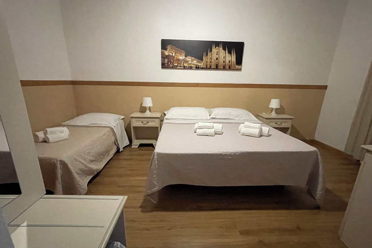 Quarto de hotel com duas camas de solteiro e uma cama de casal, cada uma com roupa de cama e toalhas brancas. Há mesas de cabeceira com abajures e um quadro com a imagem de um edifício na parede.