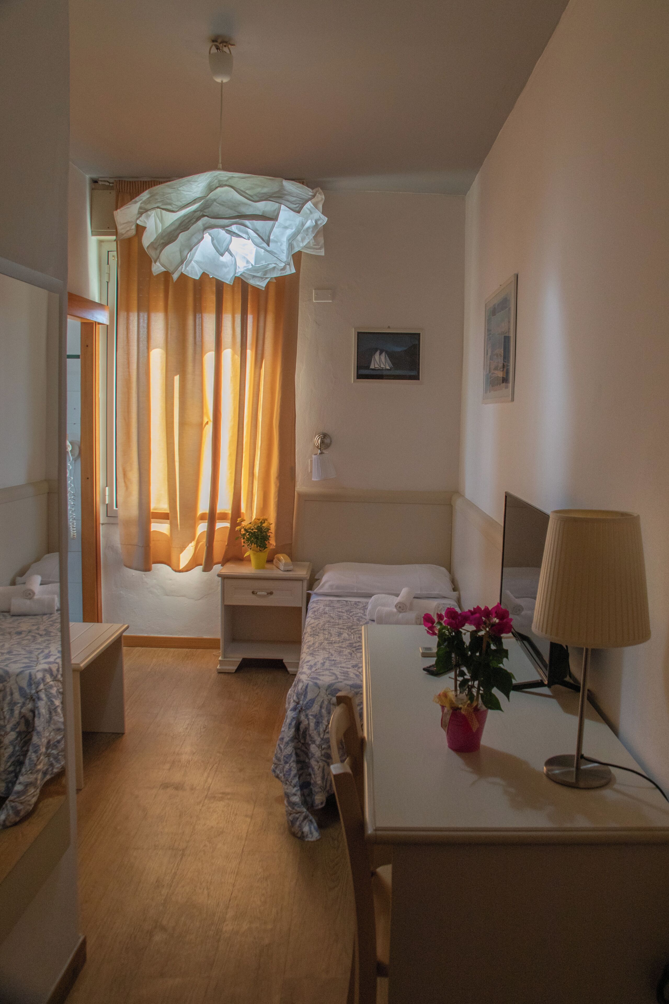 A hotel room with two beds, a desk, a chair, a lamp, a potted plant, and a window with curtains.