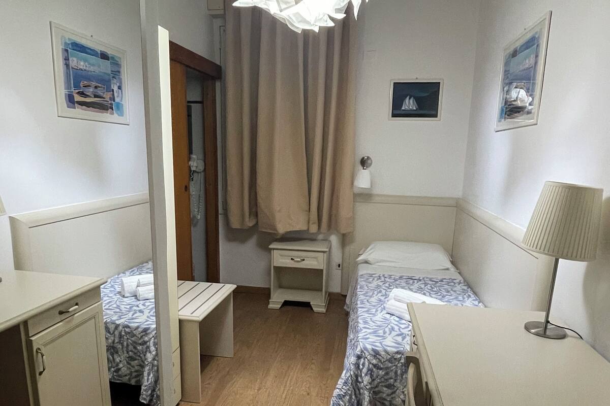 Quarto com duas camas de solteiro, mesa de cabeceira, escrivaninha, cadeira, abajur, luminária pendente e uma porta.