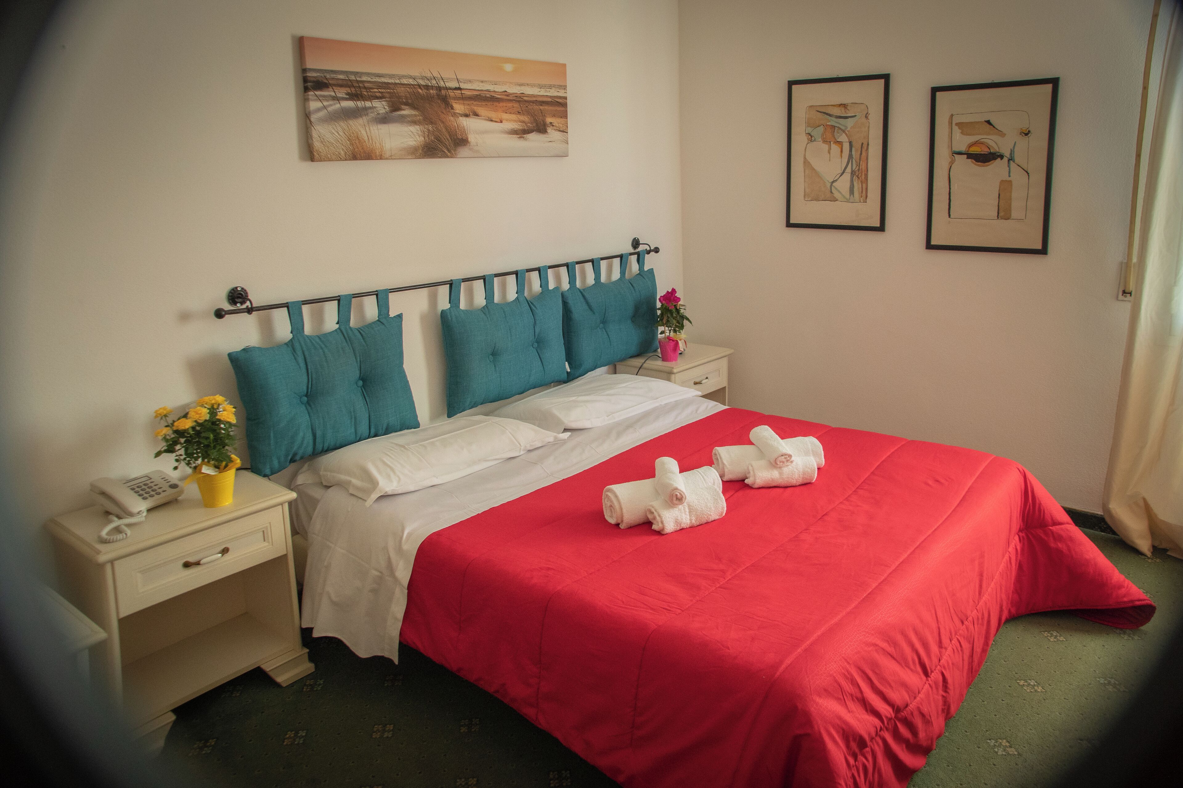 A neatly made bed with a red bedspread and white linens, a nightstand with a phone and a vase of flowers, and two framed artworks on the wall.
