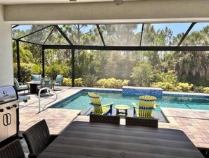 Et poolområde med en overdækket terrasse, udendørs møbler og udsigt over den omkringliggende vegetation.